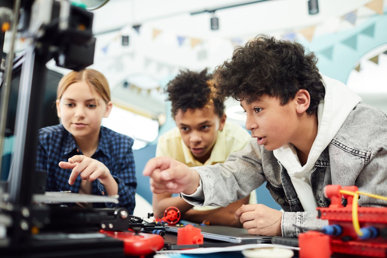 Home Three teenagers focused on a STEM project using a 3D printer in a collaborative indoor setting.